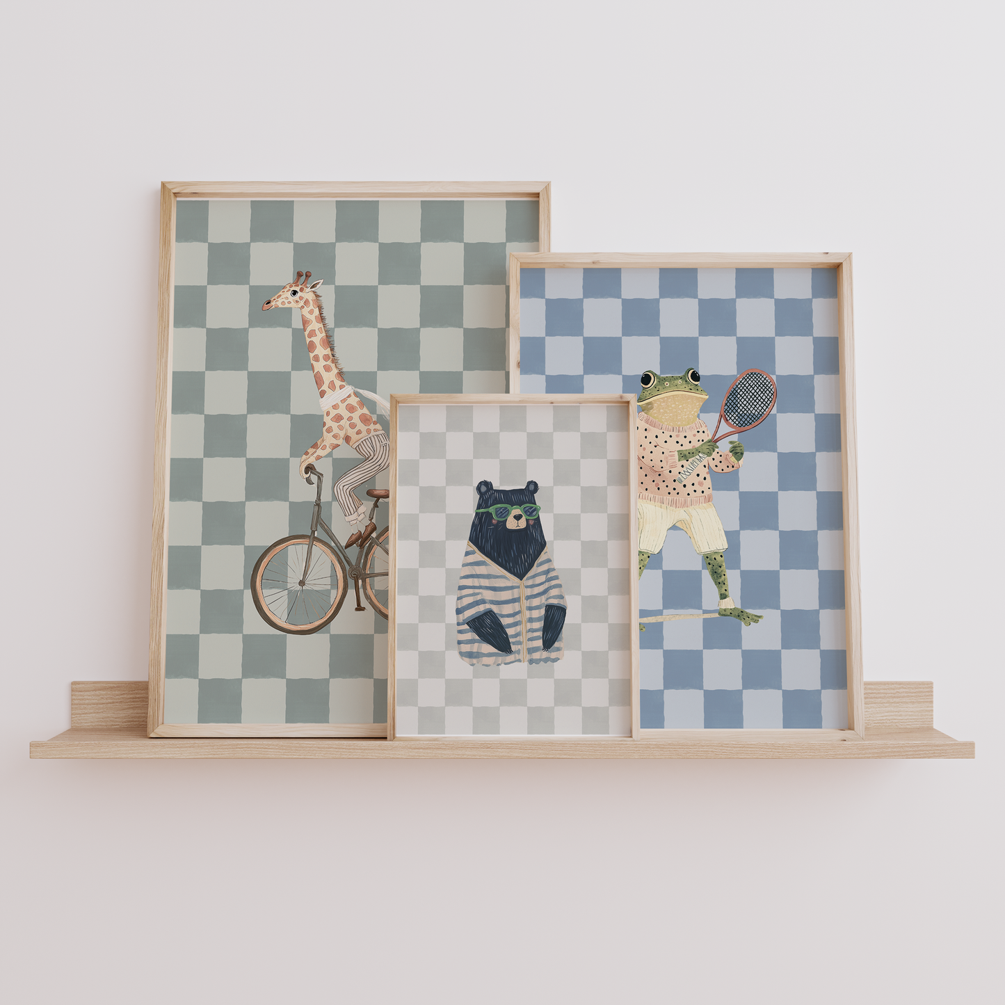 Set of three funky nursery wall art prints – giraffe on a bike, bear in sunglasses, and preppy frog playing tennis – displayed on a wooden shelf. Stylish boys’ nursery, bedroom, or playroom wall decor.