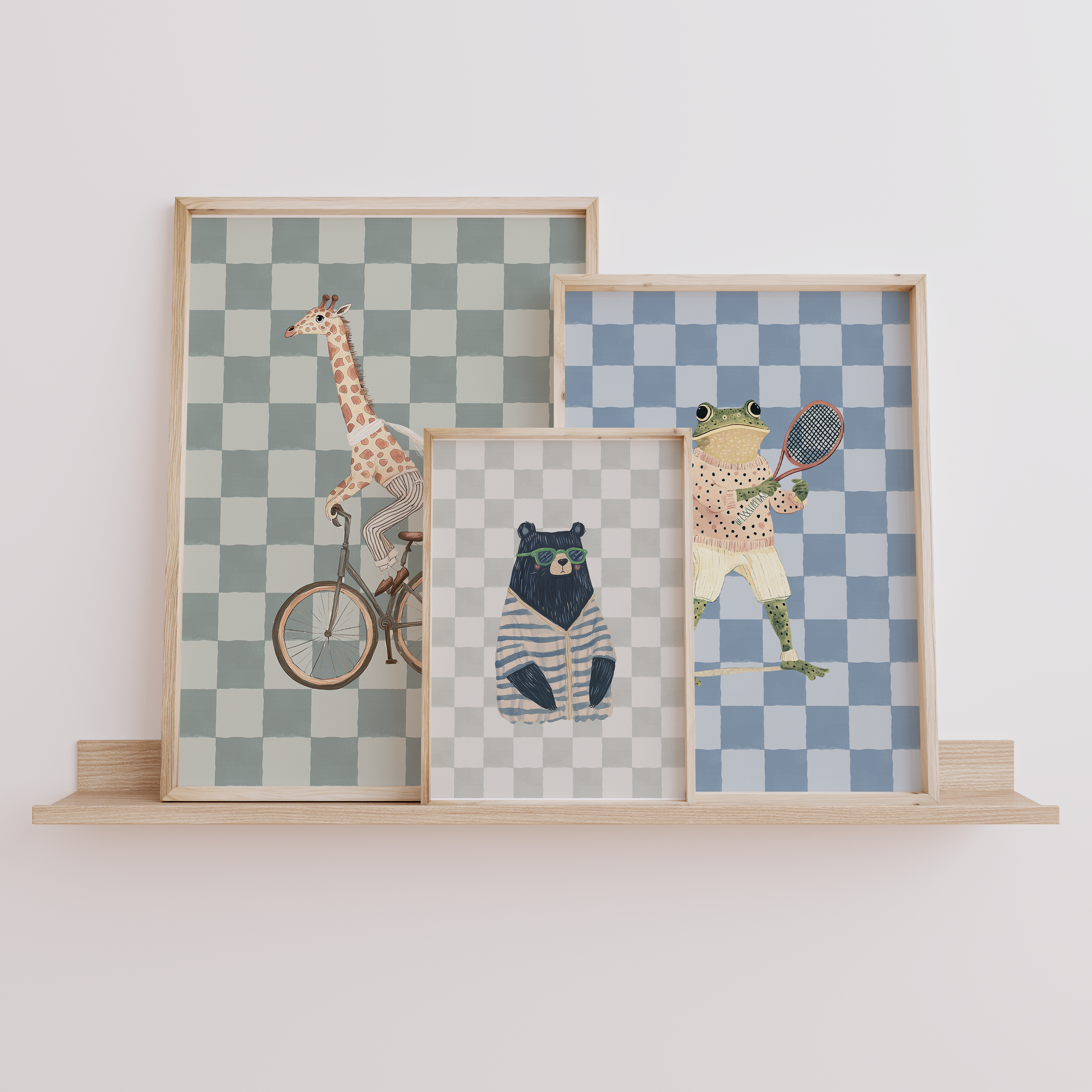 Set of three funky nursery wall art prints – giraffe on a bike, bear in sunglasses, and preppy frog playing tennis – displayed on a wooden shelf. Stylish boys’ nursery, bedroom, or playroom wall decor.