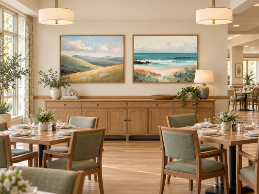 Set of 2 framed rolling hills and coastal beach wall art prints in oak frames above a wooden sideboard in a senior living dining room with a handrail and sage green dining chairs
