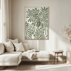 Single botanical leaf wall art print in a light wood frame styled above a cream sectional sofa, featuring soft green foliage artwork in a neutral living room setting