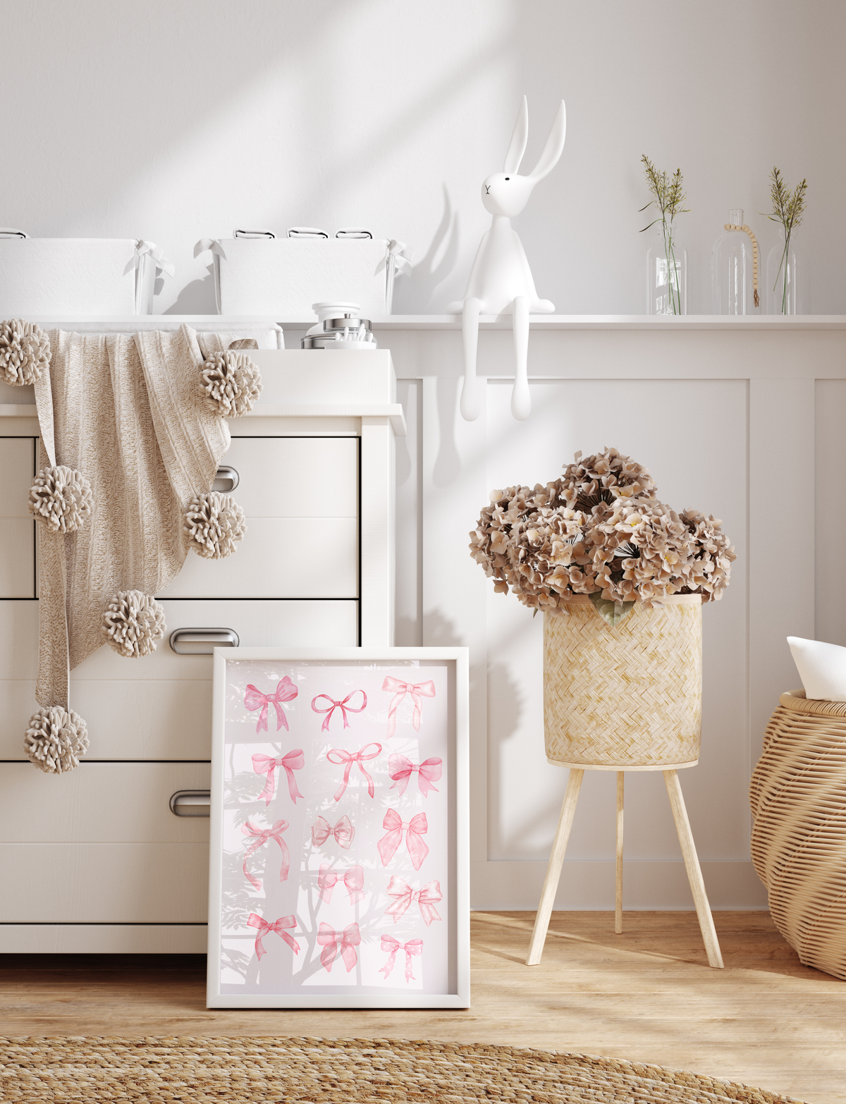 A white-framed wall art print featuring pink watercolor bows on a white background, styled in a bright, modern nursery setting next to a white dresser.