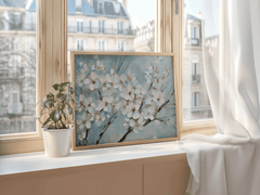 Framed cherry blossom wall art with white flowers on blue background displayed on a windowsill beside a green potted plant.