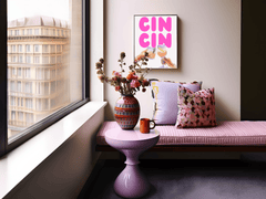 Cin cin print above a pink upholstered bench with patterned pillows beside a city window