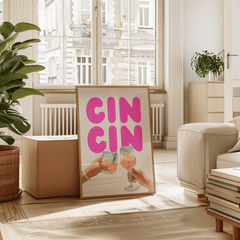 Cin cin wall art in a natural frame leaning in a sunny living room with plants and a cream armchair