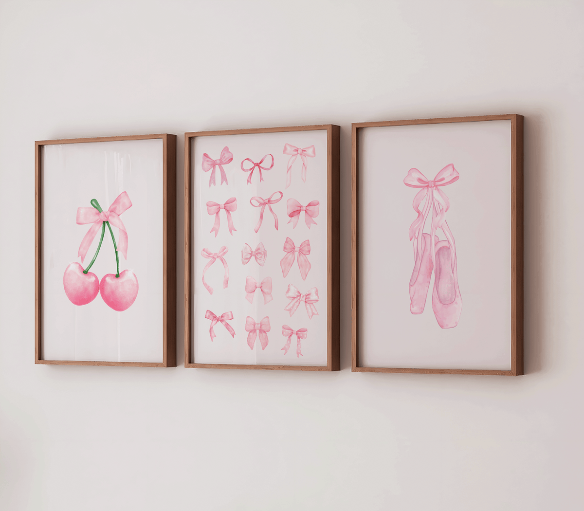 Three framed watercolor art prints with light wood frames hang on a white wall. Pink cherries tied with a bow, multiple pink bows, and pink ballet slippers with a bow.
