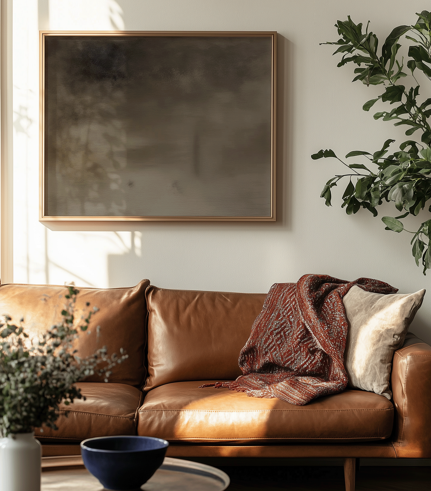 Earthy brown abstract wall art in a warm wood frame hanging above a caramel leather sofa with a textured red patterned throw blanket in a sunlit living room styled with indoor greenery.