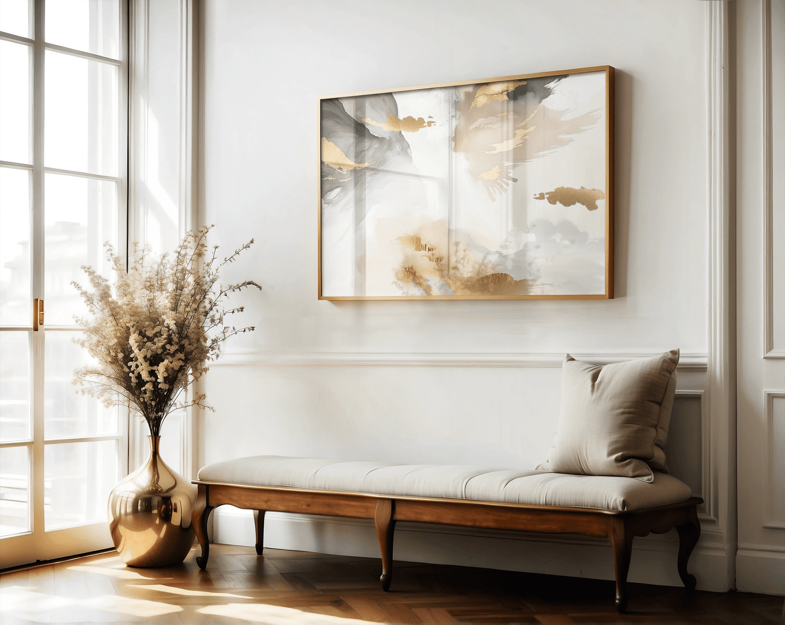 A large abstract wall art print framed with a brown wooden frame over a bench in a hallway next to a bright window and vase with dried flowers. The artwork has a mix of blush pink tones, gold accents, white, beige, light grey and charcoal.