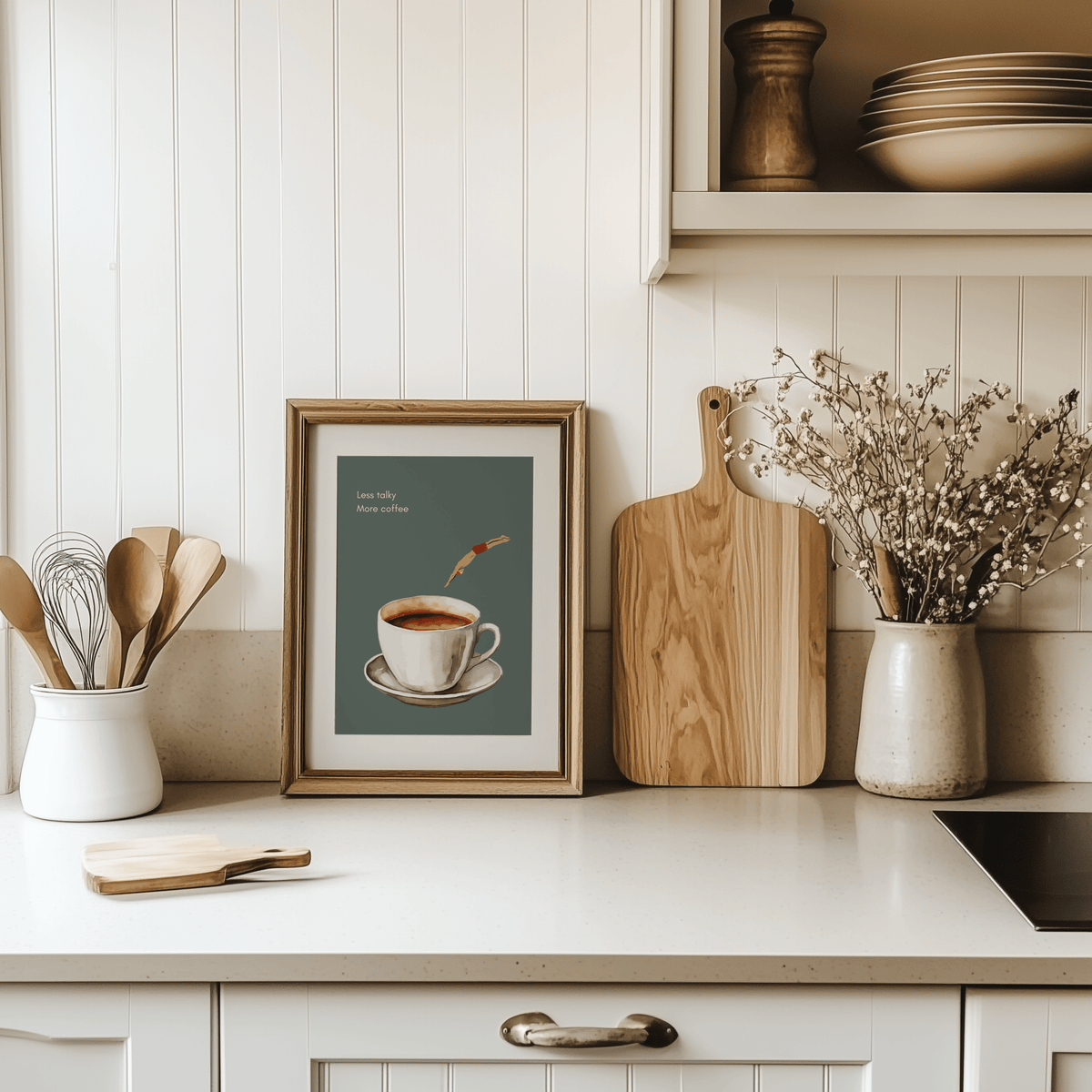 Coffee print in a natural wood frame leaning on a white kitchen counter beside a cutting board and utensils