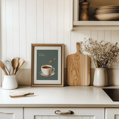 Coffee print in a natural wood frame leaning on a white kitchen counter beside a cutting board and utensils