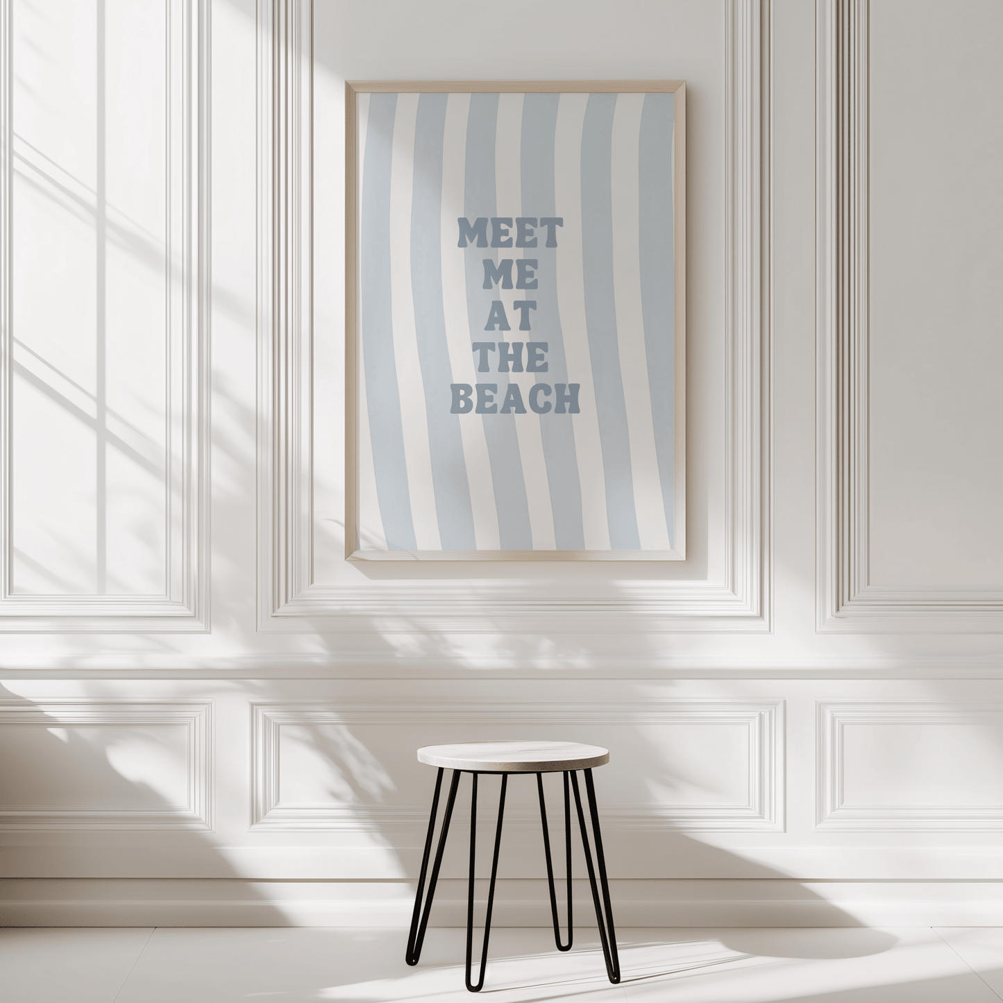Natural framed meet me at the beach wall art displayed on a white panelled corner wall with a stool in front