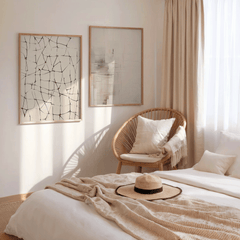 Set of two abstract wall art prints in black on beige with light wood frames styled above a bed in a neutral bedroom with woven chair, linen throw, and straw hat on blanket