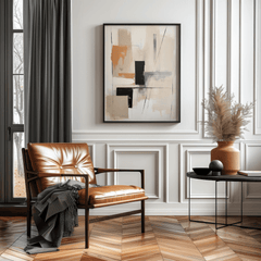 Minimalist tan and black geometric canvas print in a black frame styled over a leather accent chair in a classic paneled corner.