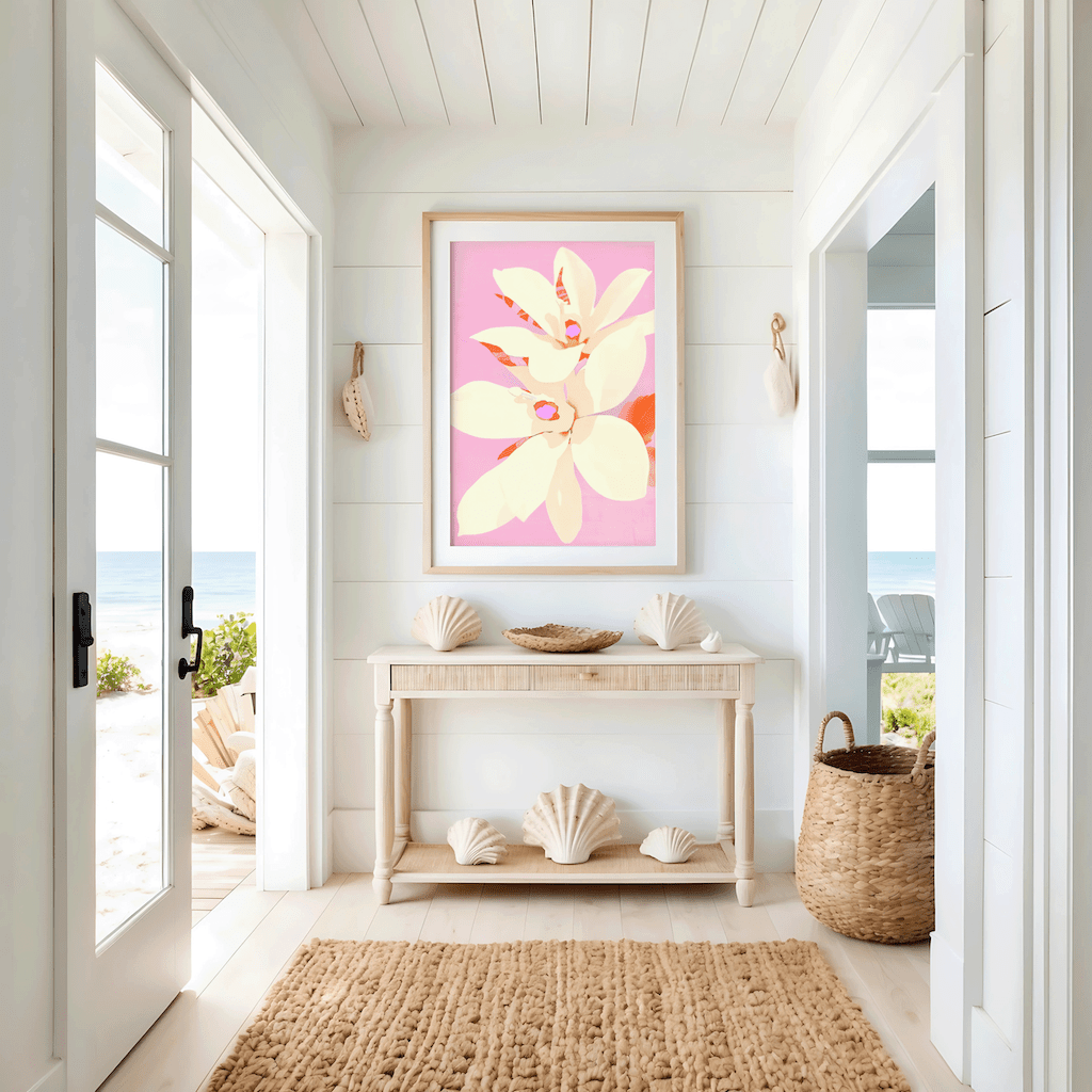 A pale yellow or ivory floral wall art print framed with coral stems and blush background in a modern botanical illustration style. The artwork is displayed in a modern beach house entryway above a light wooden console table with white walls, windows, natural light and oceanview.