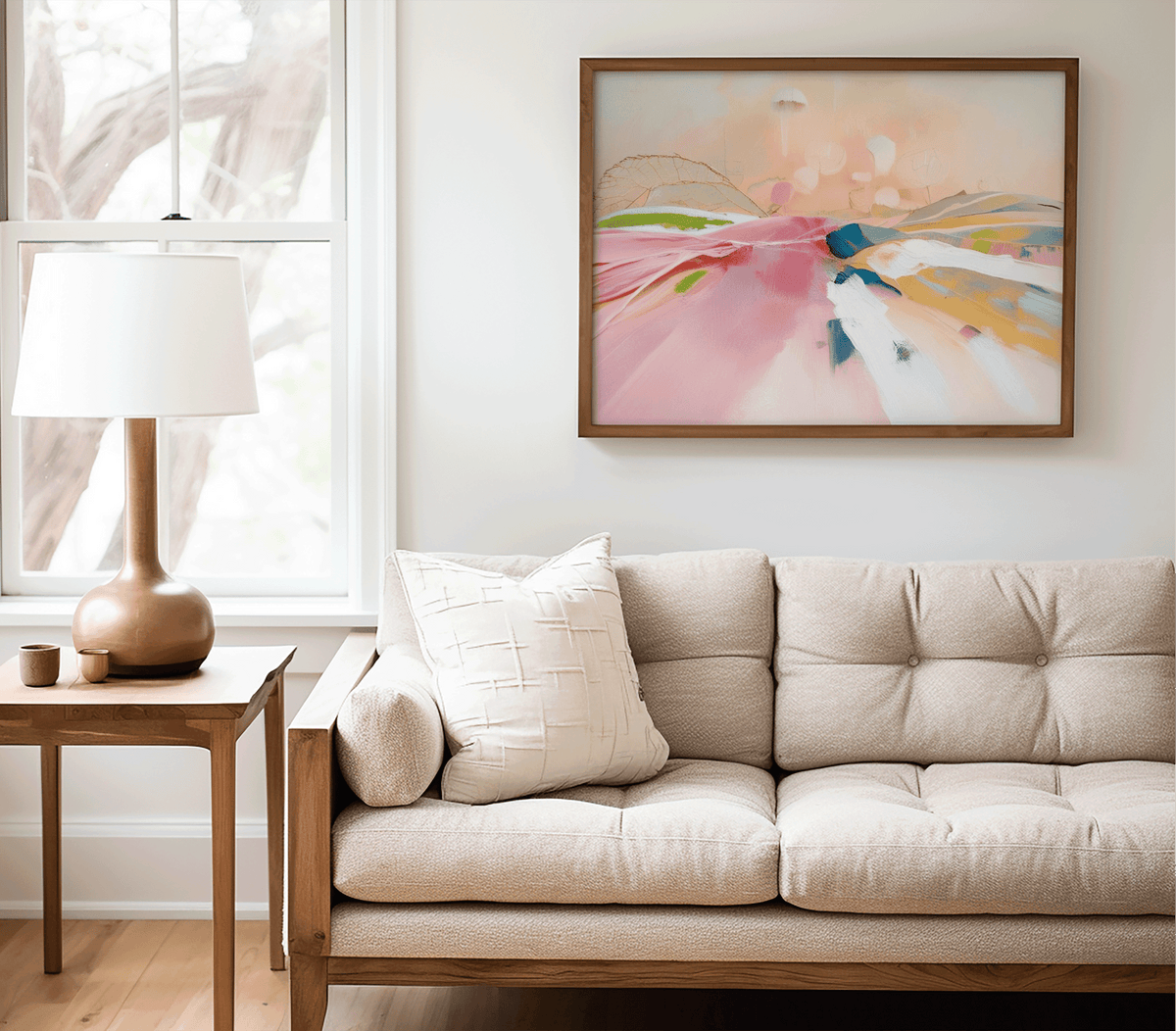 Pastel pink abstract landscape print in a natural wood frame above a beige tufted sofa with a table lamp in a neutral living room