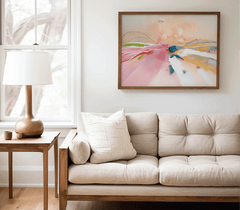 Pastel pink abstract landscape print in a natural wood frame above a beige tufted sofa with a table lamp in a neutral living room