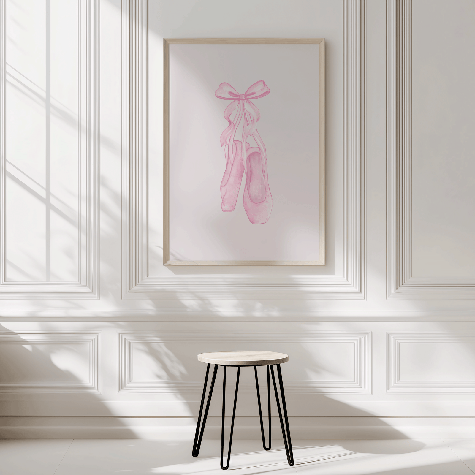 Framed artwork of pink ballet shoes with a bow on a white wall with wainscoting.