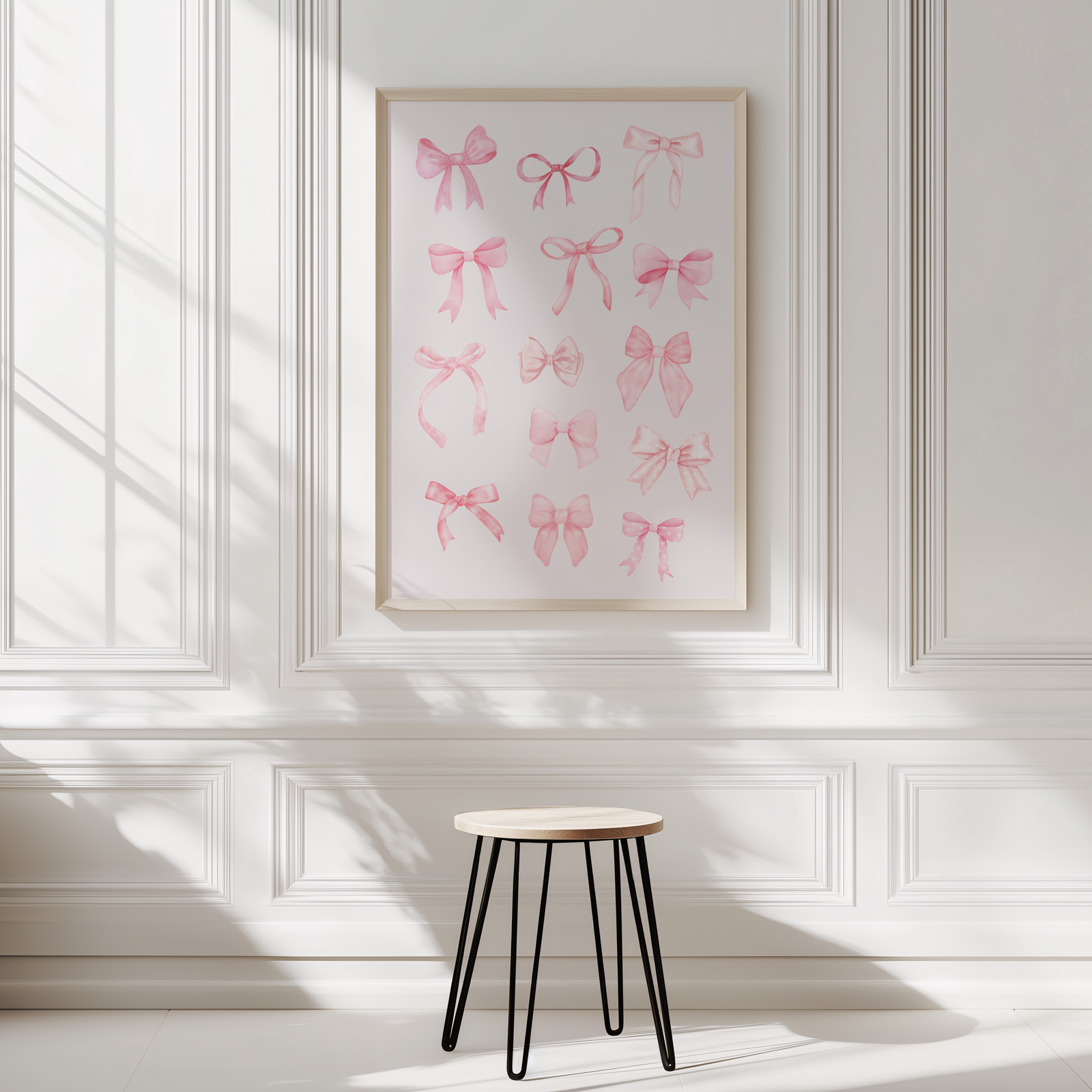 A framed wall art print featuring pink watercolor bow illustrations, displayed on a white panelled wall above a small stool