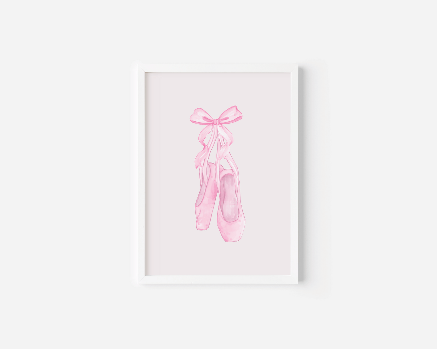 A white-framed canvas wall art print featuring a watercolor illustration of pink ballet pointe shoes tied with a bow, displayed against a white wall.