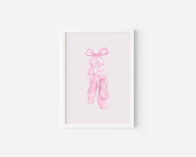 A white-framed canvas wall art print featuring a watercolor illustration of pink ballet pointe shoes tied with a bow, displayed against a white wall.