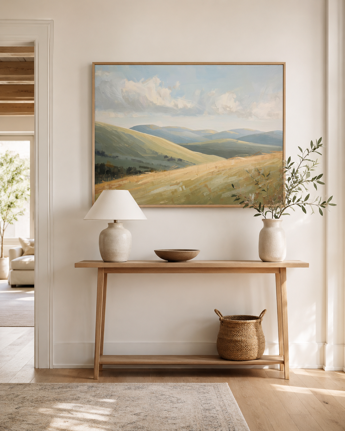 A framed nature landscape canvas wall art print of rolling green and beige hills under a cloudy sky, on a wall above a light wood console table in entryway.