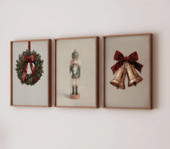 Three framed Christmas prints on a neutral wall featuring a plaid ribbon wreath, a vintage navy-and-gold nutcracker, and a pair of gold bells tied with a deep red velvet bow