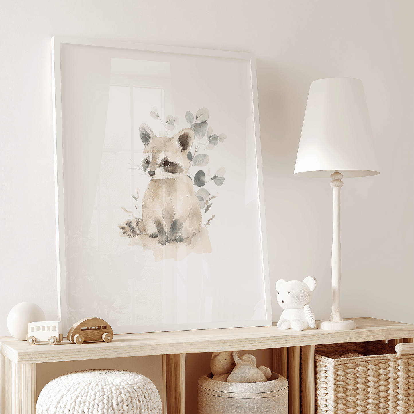 Neutral minimal woodland nursery animal art of a baby raccoon in a watercolor style with soft browns, beiges, and greens with eucalyptus leaves in a white frame on a wooden shelf in a kid's play room.